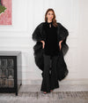 A person wearing a black pleated organza stole with a wide neckline and long sleeves, paired with black trousers in a luxurious setting.